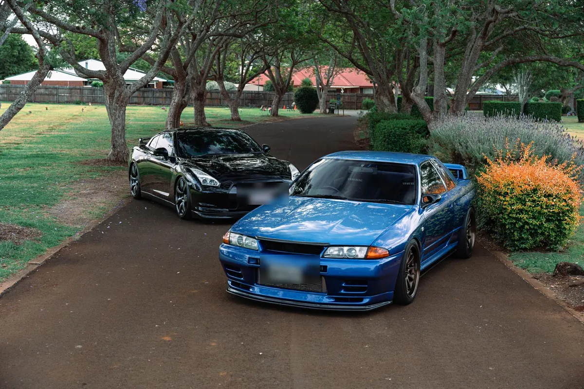 Black R35 GT-R and blue R32 GT-R detailed side by side under jacaranda trees